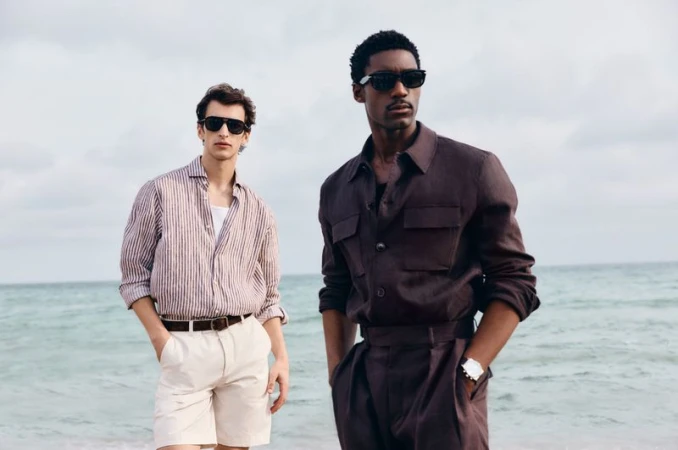 Two adult men pose on a beach wearing casual summer attire, including button-down shirts, shorts, trousers, and sunglasses.