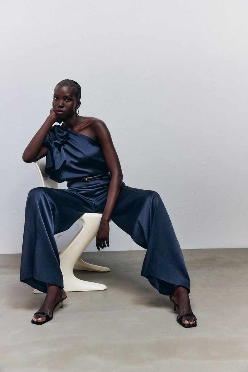 Dark-skinned adult female in navy, one-shoulder satin jumpsuit sitting on a chair in front of a white wall, shot in photo studio.