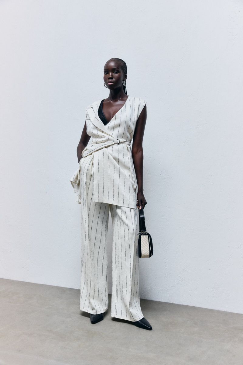 Editorial fashion shot of a Black female model posing in a beige and black striped pantsuit with a shoulder bag against a white background.