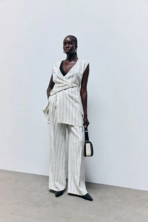 Editorial fashion shot of a Black female model posing in a beige and black striped pantsuit with a shoulder bag against a white background.