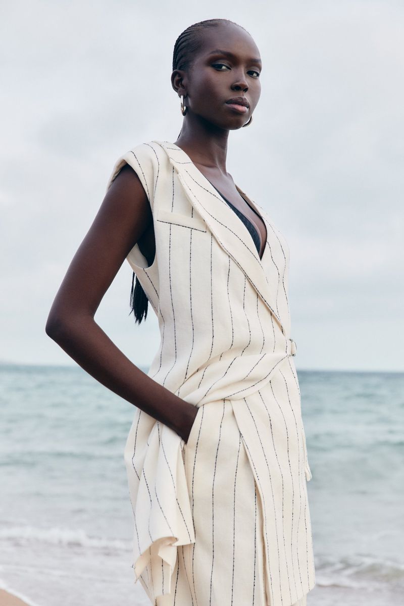 Fashion shot of a Black woman in a pinstripe dress on a beach with neutral sky and water backdrop, fashion, style, and outdoor photoshoot.
