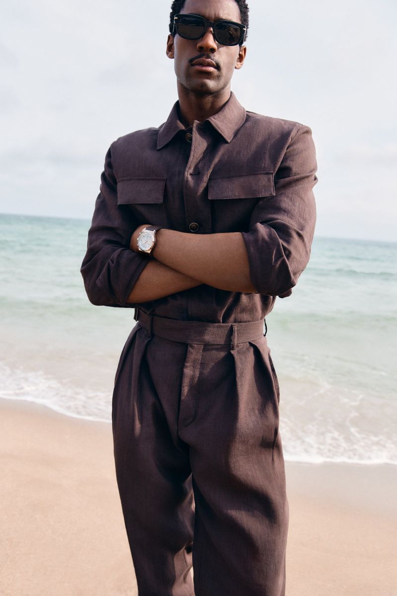 Model wearing dark sunglasses and a brown matching shirt and trousers standing with arms crossed on a sandy beach.