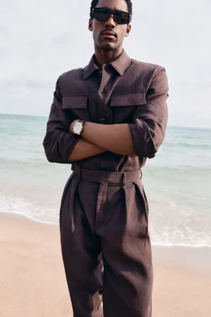 Model wearing dark sunglasses and a brown matching shirt and trousers standing with arms crossed on a sandy beach.