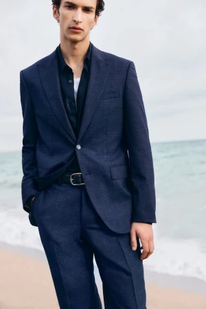 An adult man in a navy suit and black shirt poses on a beach with tan sand and ocean water on a cloudy day, fashionable and stylish.