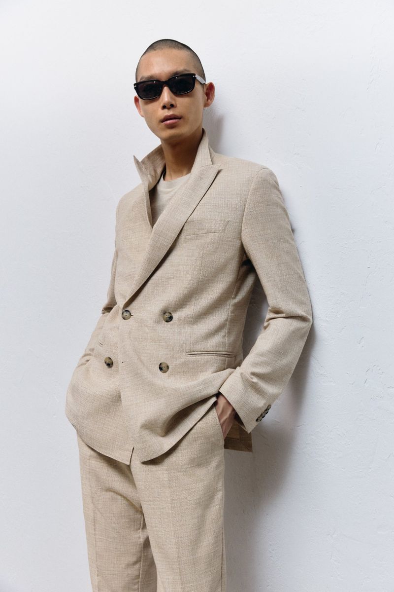A slender male fashion model wearing a beige-colored suit and black sunglasses poses in front of a white wall indoors.