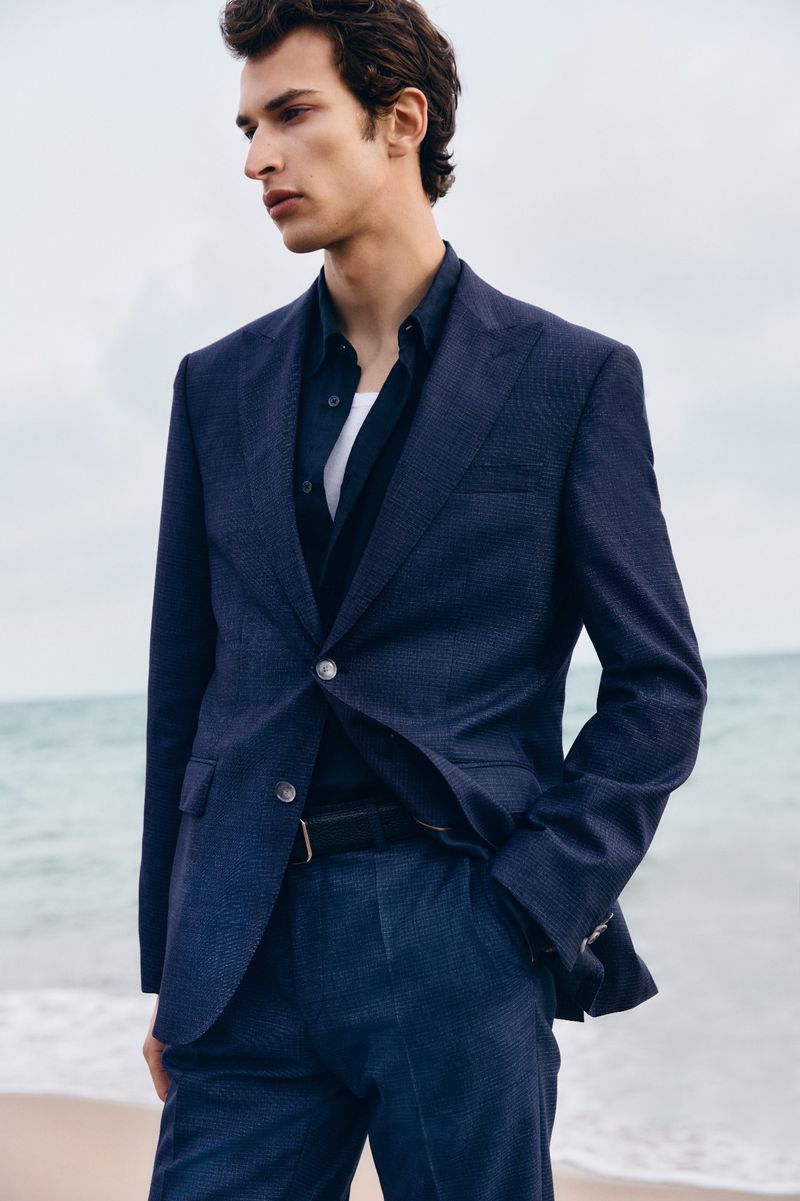 Man wearing a navy blue blazer and matching trousers standing outdoors against a beach background