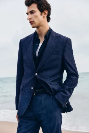 Man wearing a navy blue blazer and matching trousers standing outdoors against a beach background