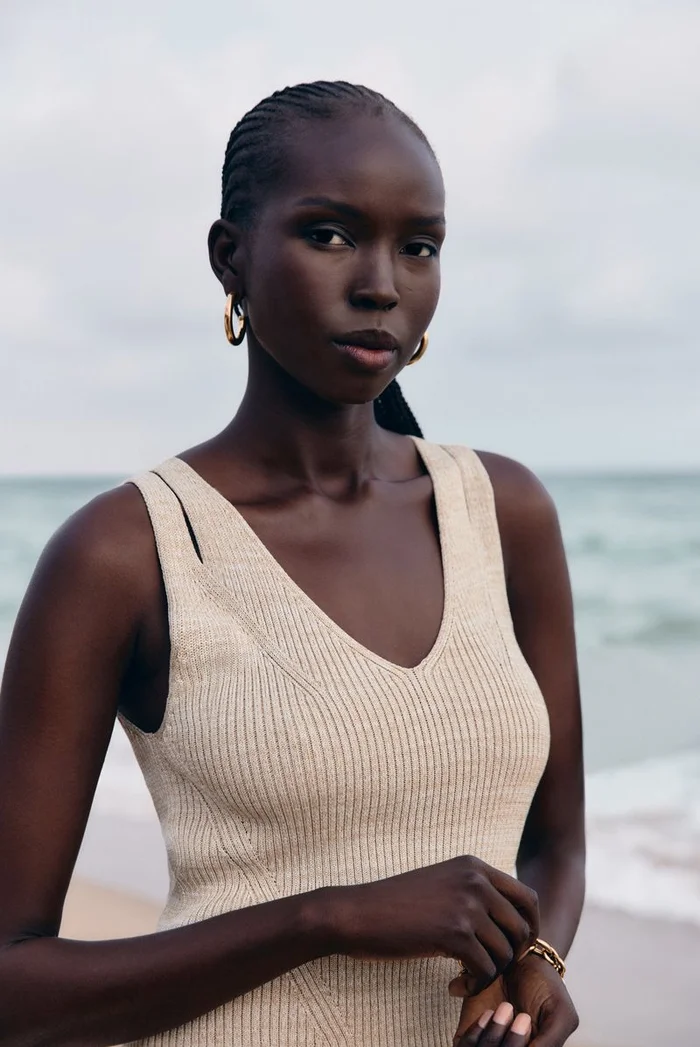 Model Ajok Madel wearing a cream sleeveless ribbed dress by BOSS against an ocean beach backdrop