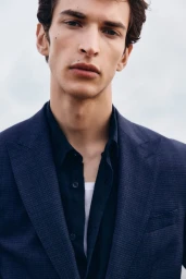Close-up portrait of a male model wearing a navy blazer and dark button-down shirt against an overcast sky.