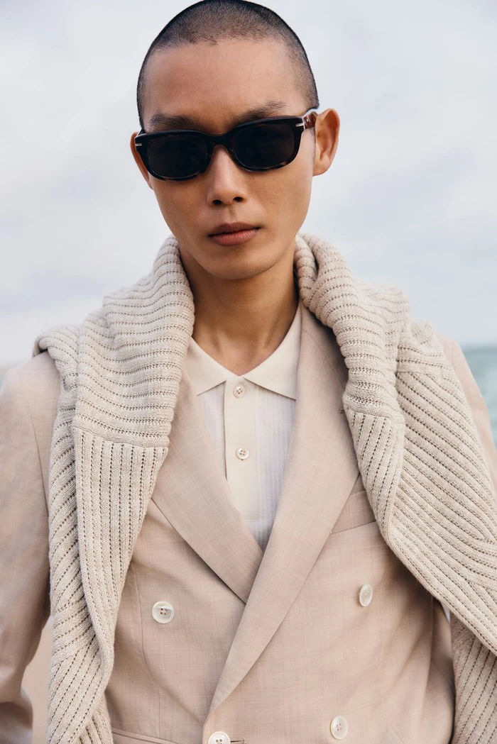 Model wearing dark sunglasses, beige double-breasted suit jacket, and cream sweater draped over shoulders outdoors