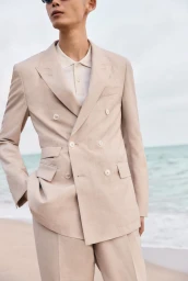 Model wearing a beige double-breasted suit jacket standing against an ocean beach background.
