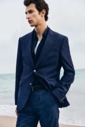 Man wearing a navy blue blazer and matching trousers standing outdoors against a beach background