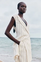 Model Ajok Madel wearing a cream sleeveless pinstripe vest by BOSS against an ocean backdrop