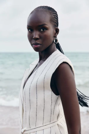 Model Ajok Madel wearing a cream sleeveless pinstripe vest by BOSS against an ocean backdrop for SS25 Occasion.