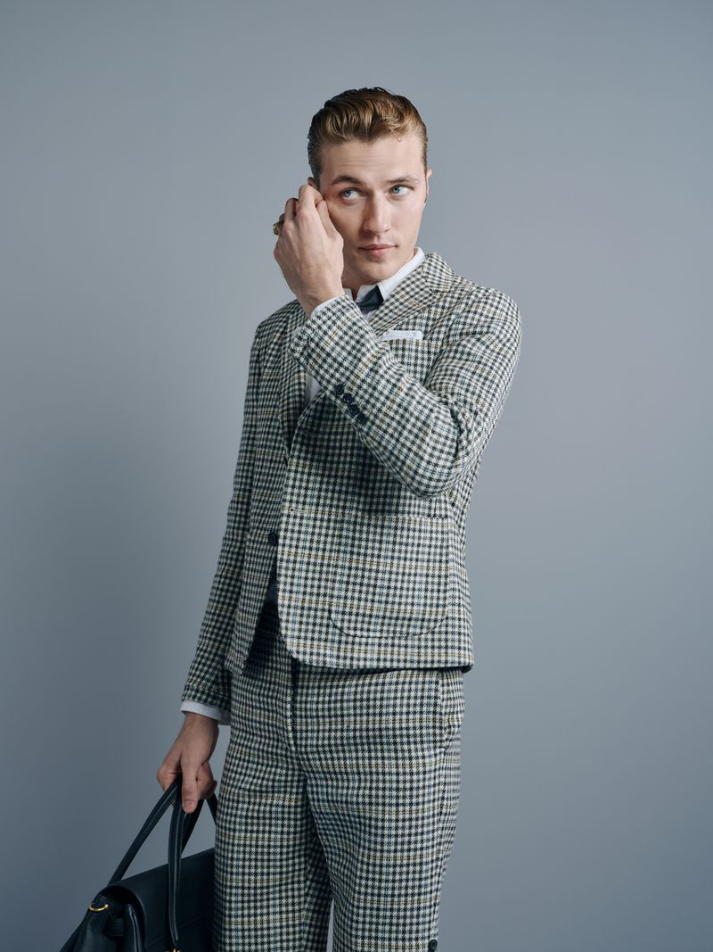 Man in a patterned suit holds a black bag, looking up in a studio setting with soft lighting and a neutral gray background.