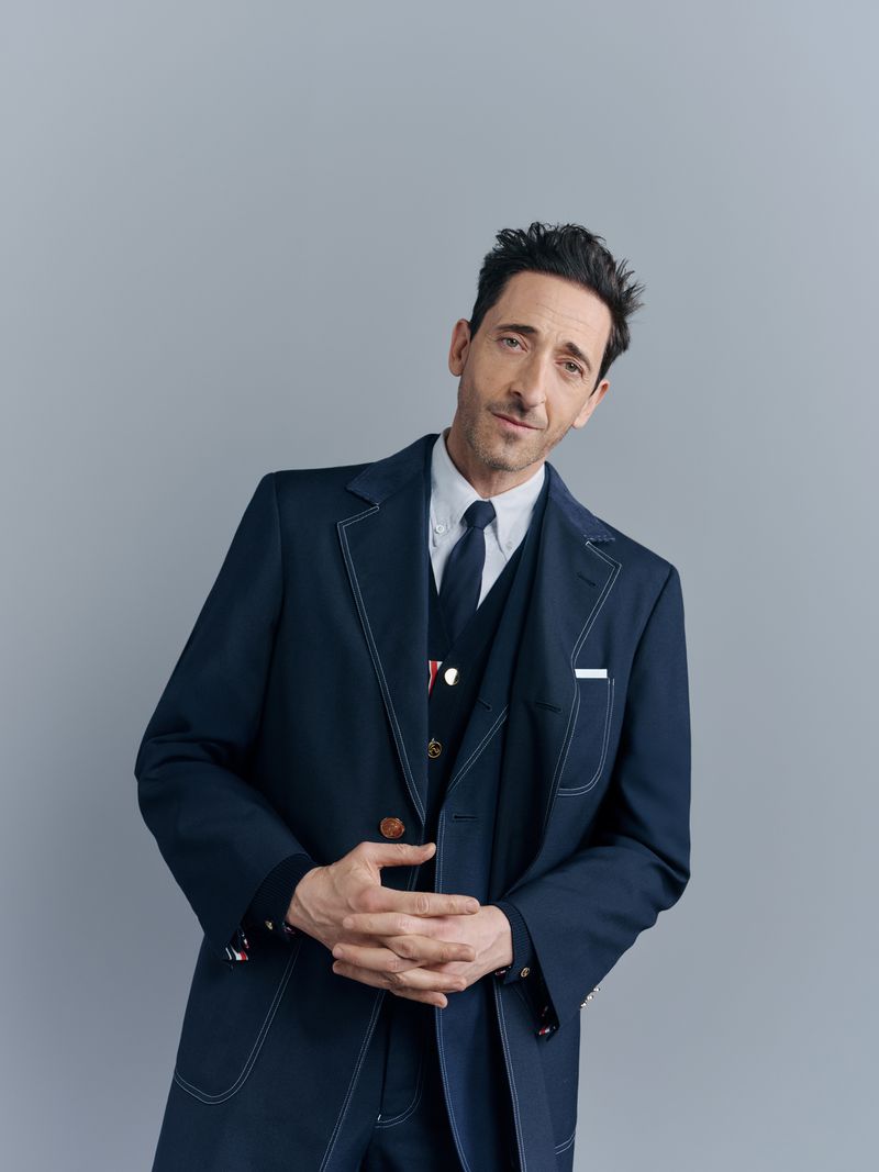 Portrait of adult male in dark blue suit with tie and vest against light gray backdrop looking at camera with hands clasped.