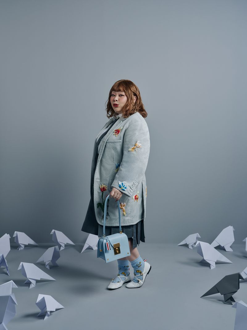 Woman in light blue coat, holding purse, poses amid paper origami birds in a studio with soft lighting and a pale gray background.