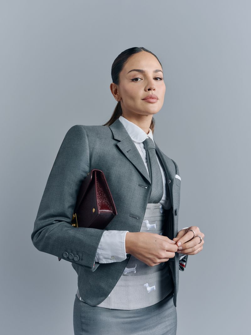 Portrait of a brunette woman in grey suit and tie holding a burgundy clutch against grey backdrop, looking at the camera.