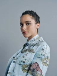 Portrait of a woman wearing a light blue blazer with a scenic landscape print against a neutral grey background
