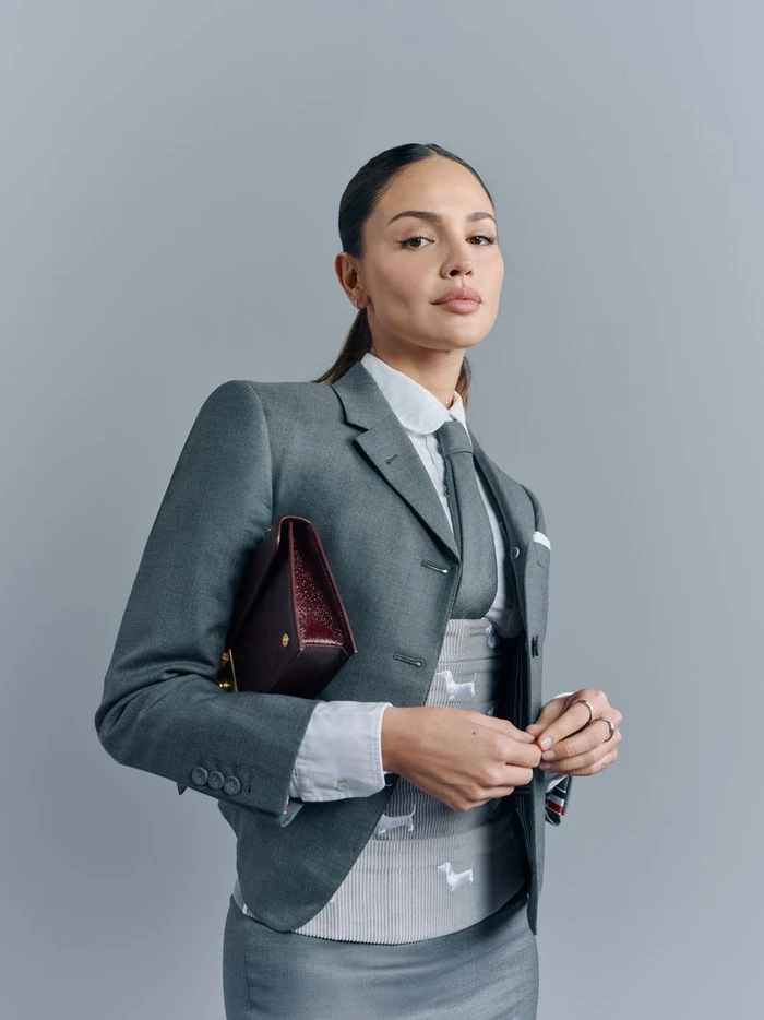 Eiza Gonzalez in a grey Thom Browne suit holding a dark handbag against a solid grey background
