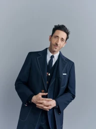 Adrien Brody wearing a tailored navy suit with white contrast stitching against a plain grey studio backdrop for Thom Browne FW25.