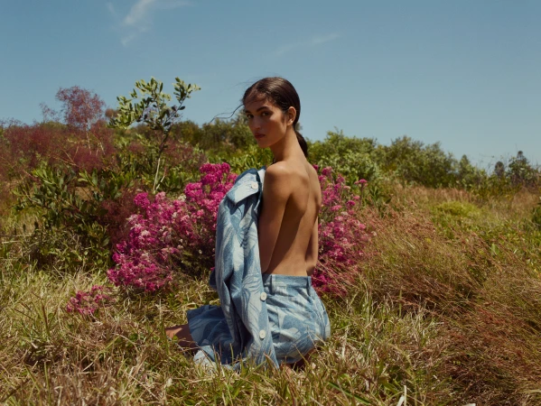 A woman crouching in a grassy meadow with vibrant pink wildflowers, wearing a patterned blue jacket and matching trousers.