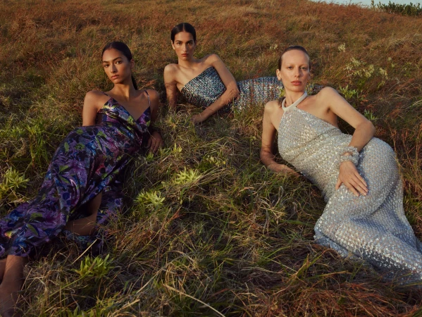 Three female models in evening dresses lying in a grassy field outdoors under natural sunlight.