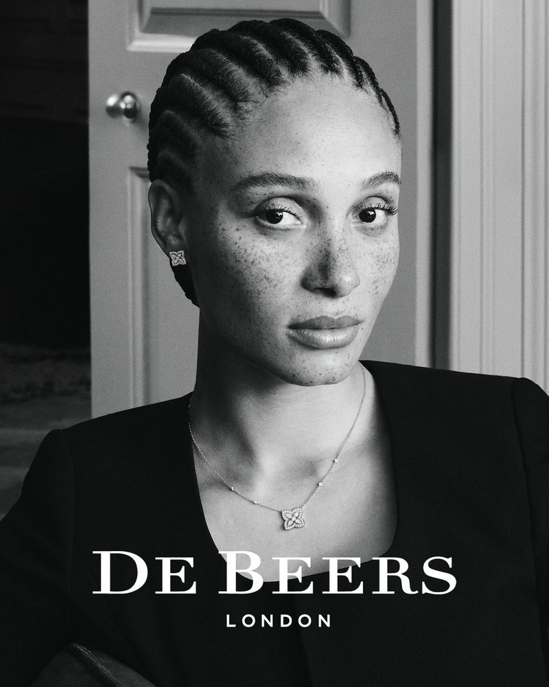 Portrait of a Black female adult wearing stud earrings and a necklace against a muted background with the De Beers logo.