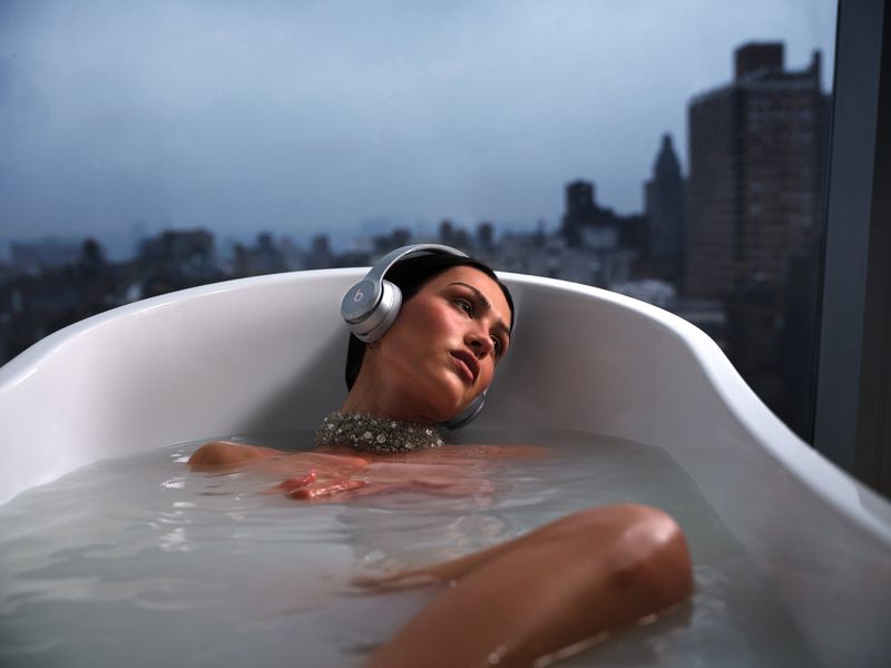 Adult female in a white tub with milky water, wearing headphones and jewelry in an indoor apartment setting with a city view.