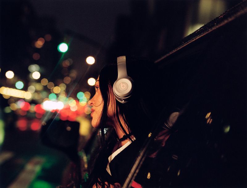 Profile view of a young adult female wearing headphones in a dark, bokeh-lit urban environment at night, car window frame visible.