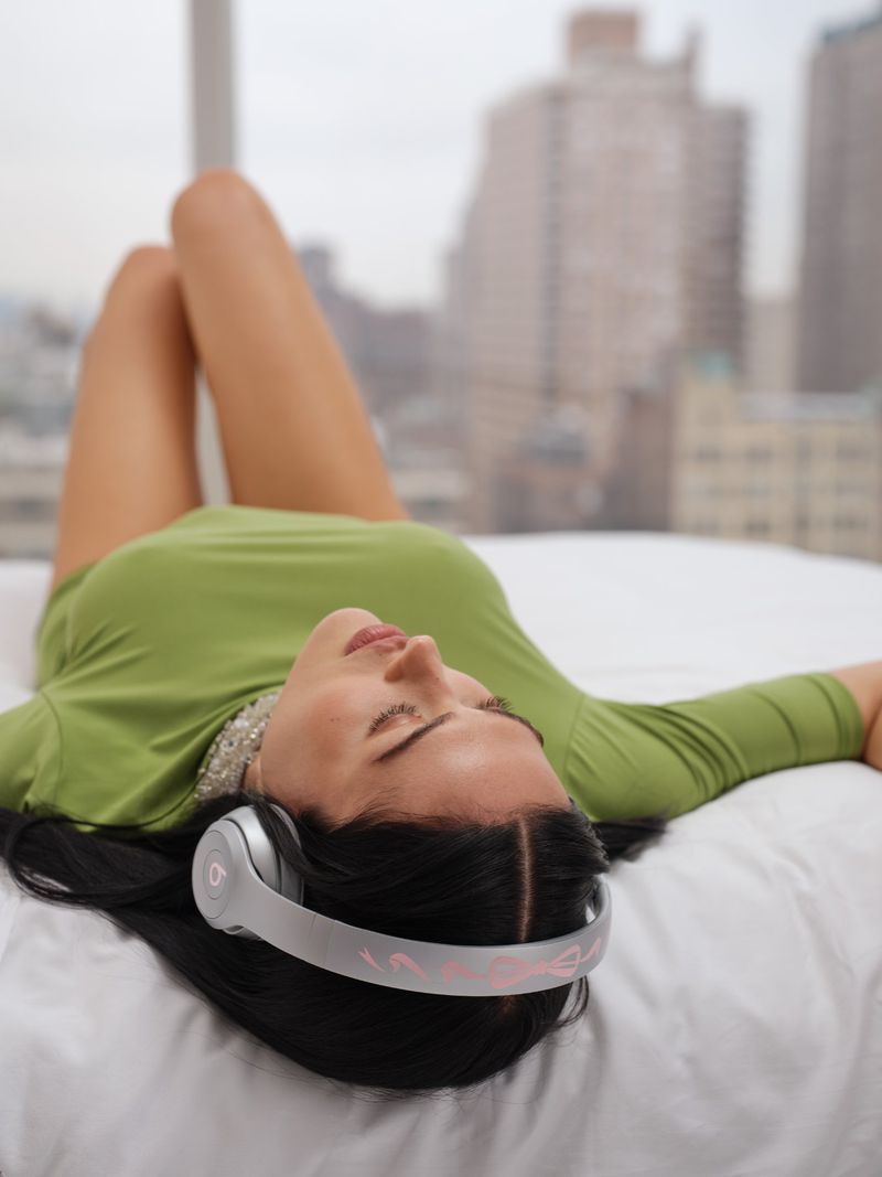 A woman with headphones lies on a bed in front of a window with a city view, wearing a green shirt, dark hair. Relaxing indoors.