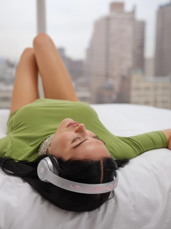 A woman with headphones lies on a bed in front of a window with a city view, wearing a green shirt, dark hair. Relaxing indoors.