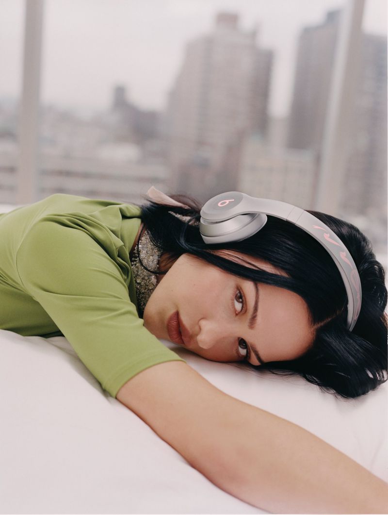 Adult female in a green top laying on a white bed wearing light grey Beats headphones with a city skyline visible in the background.