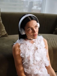 Amelia Gray wearing white wireless headphones and a textured white floral top while seated on a green sofa