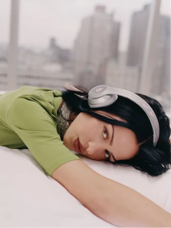 Amelia Gray wearing wireless headphones and a green top lying down indoors against a blurred city window view.
