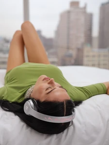 Model Amelia Gray lying on a white bed wearing a green top and silver headphones with a city background for Beats by Dre x Sandy Liang.