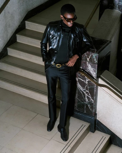 A stylish adult male in a black leather jacket and sunglasses poses on marble stairs, conveying a fashion-forward and sophisticated look.