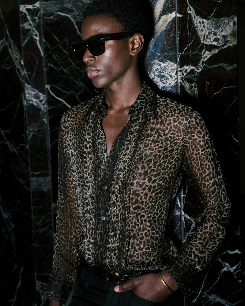 An adult male model wearing a sheer leopard-print shirt and sunglasses poses against a dark marble wall in a close-up studio portrait.