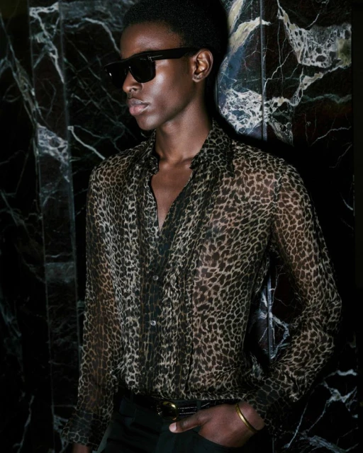 An adult male model wearing a sheer leopard-print shirt and sunglasses poses against a dark marble wall in a close-up studio portrait.