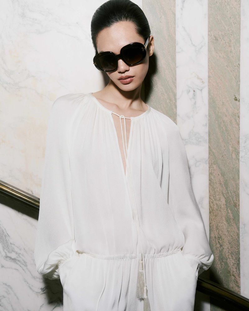Elegant woman in a white jumpsuit and sunglasses poses against a marble wall, capturing a sophisticated fashion statement with natural light.
