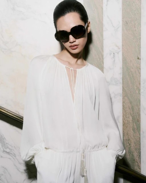 Elegant woman in a white jumpsuit and sunglasses poses against a marble wall, capturing a sophisticated fashion statement with natural light.