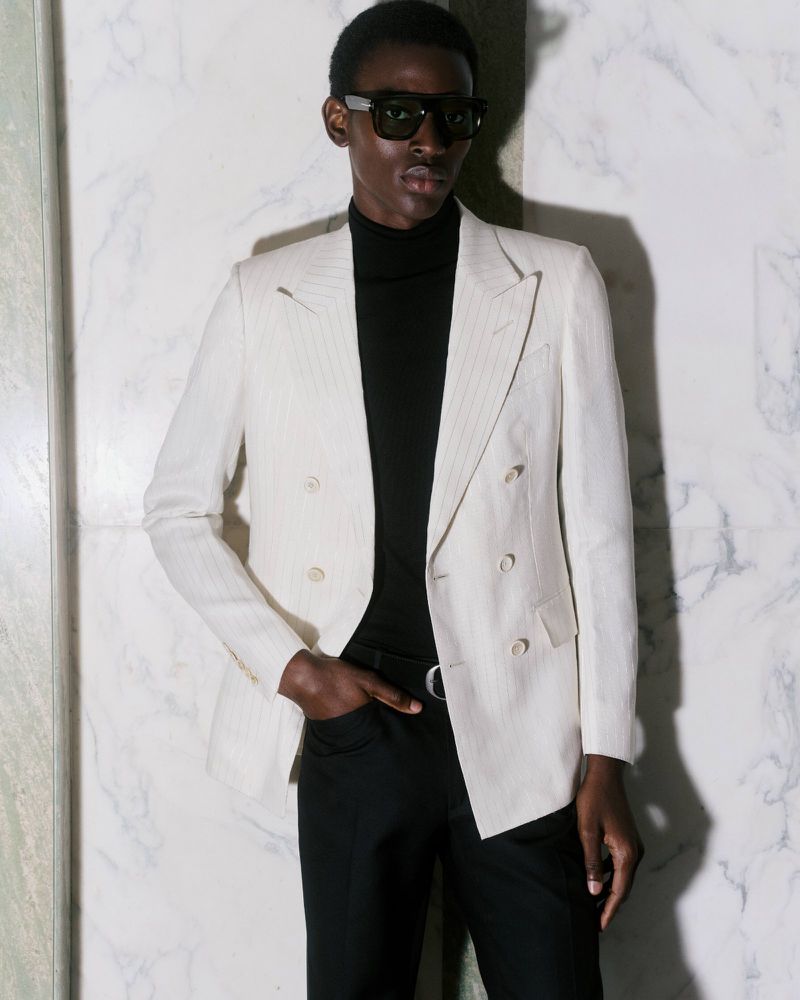 Elegant adult male poses in a double-breasted blazer, black turtleneck, and sunglasses against a mottled marble backdrop, indoor fashion.