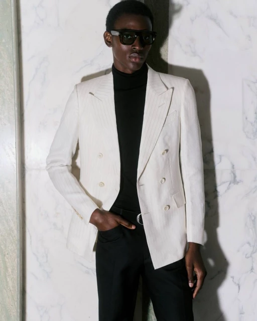 Elegant adult male poses in a double-breasted blazer, black turtleneck, and sunglasses against a mottled marble backdrop, indoor fashion.