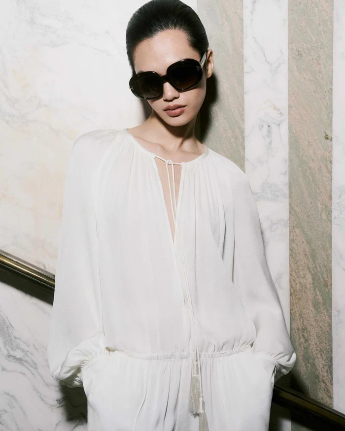 Model wearing dark sunglasses and a white long-sleeve top against a marble-patterned wall for Tom Ford SS25