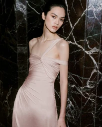 Model in a pale pink draped evening dress posing against a dark marble wall for Net-A-Porter