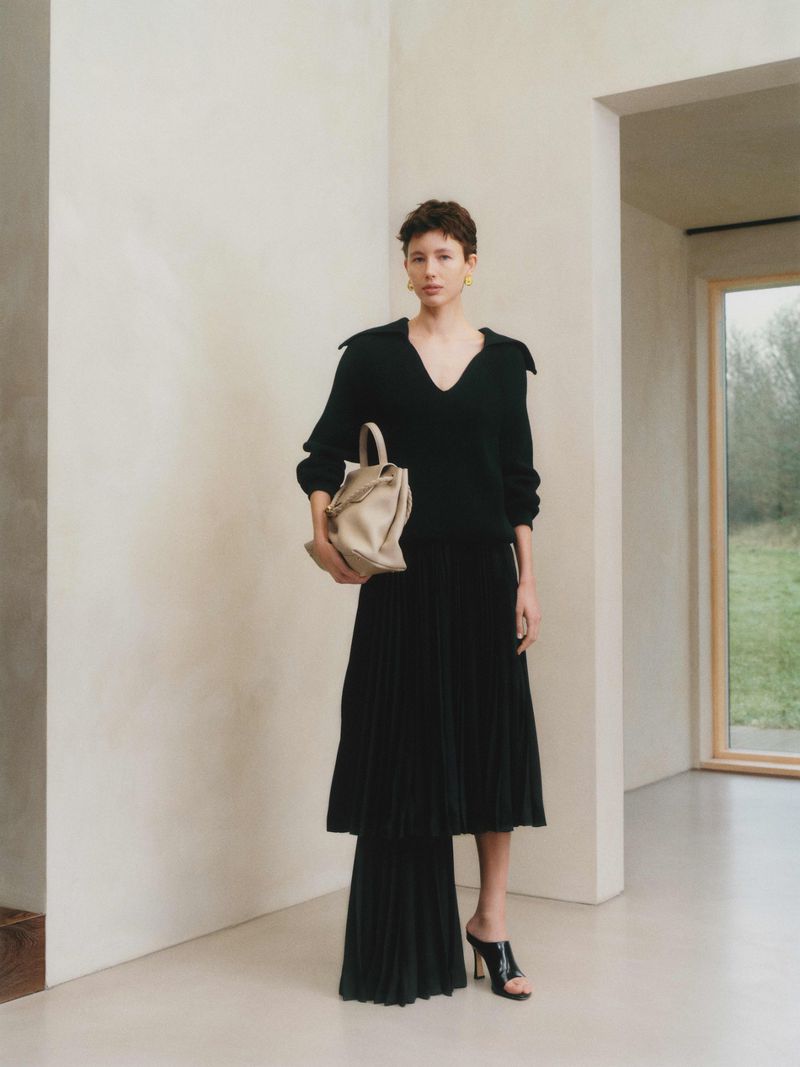 A woman in a black sweater and pleated skirt poses indoors with a tan handbag in front of a beige wall with a window.