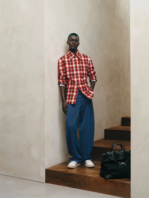 A full shot of a Black male model in a red and white checkered shirt with blue pants leaning against a beige wall next to stairs.