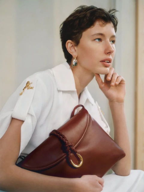 A woman with short hair poses indoors, holding a large, dark brown leather handbag with gold ring hardware and wearing gold earrings.