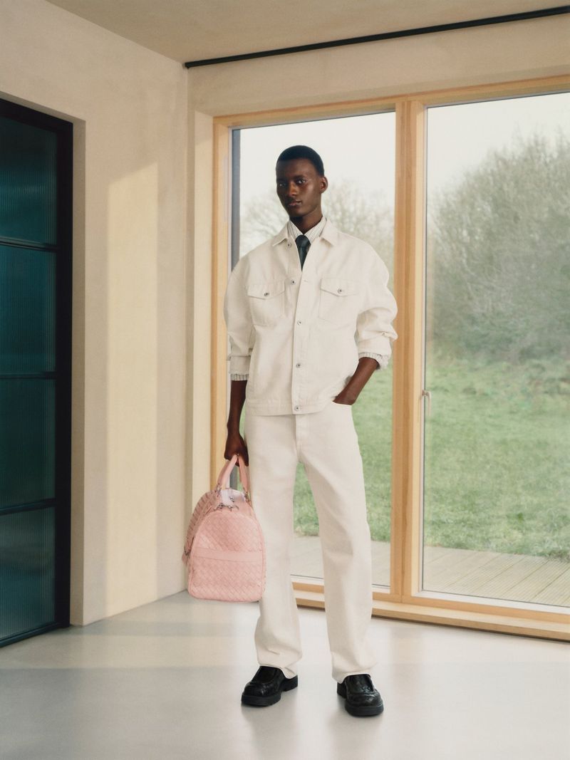 A stylish dark-skinned man in white denim stands indoors with a pink duffel bag near a window, showcasing modern fashion.