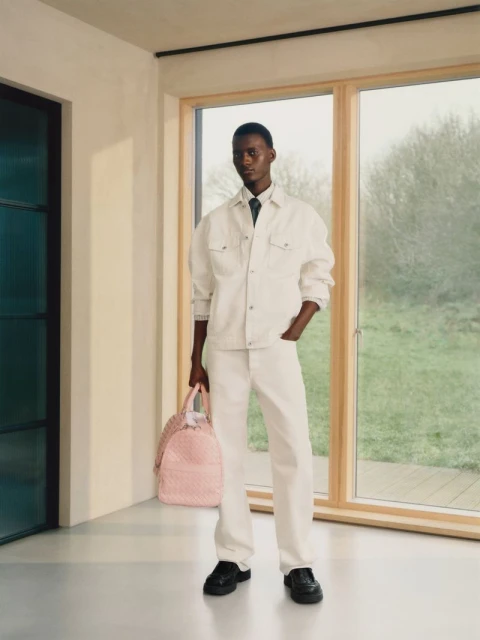A stylish dark-skinned man in white denim stands indoors with a pink duffel bag near a window, showcasing modern fashion.